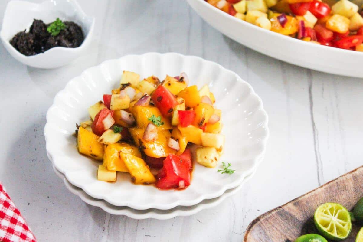 A white plate with a colorful fruit and vegetable salad containing mango, apple, tomato, and onion. A small dish of dark dip and a larger bowl of salad are in the background.