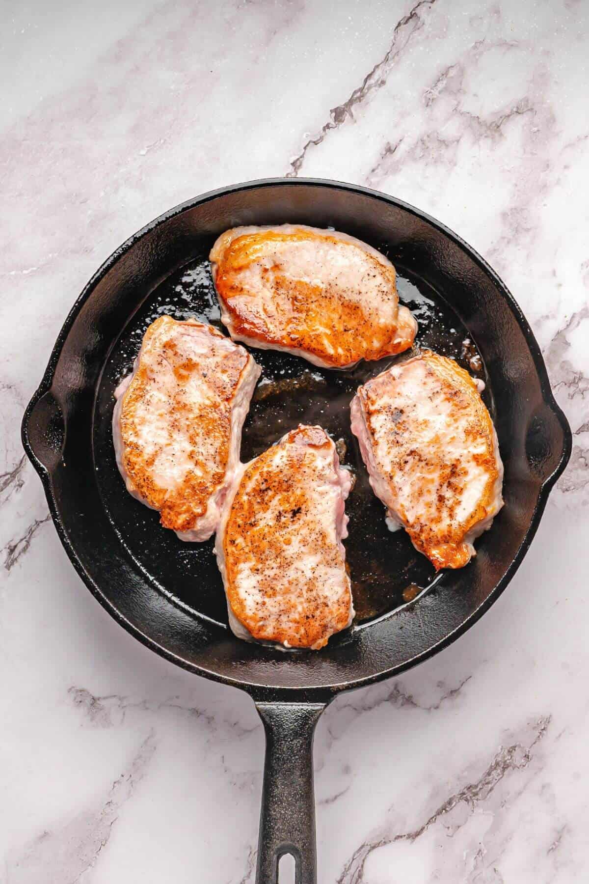 Four seasoned pork chops being seared in a black cast-iron skillet on a marble countertop.
