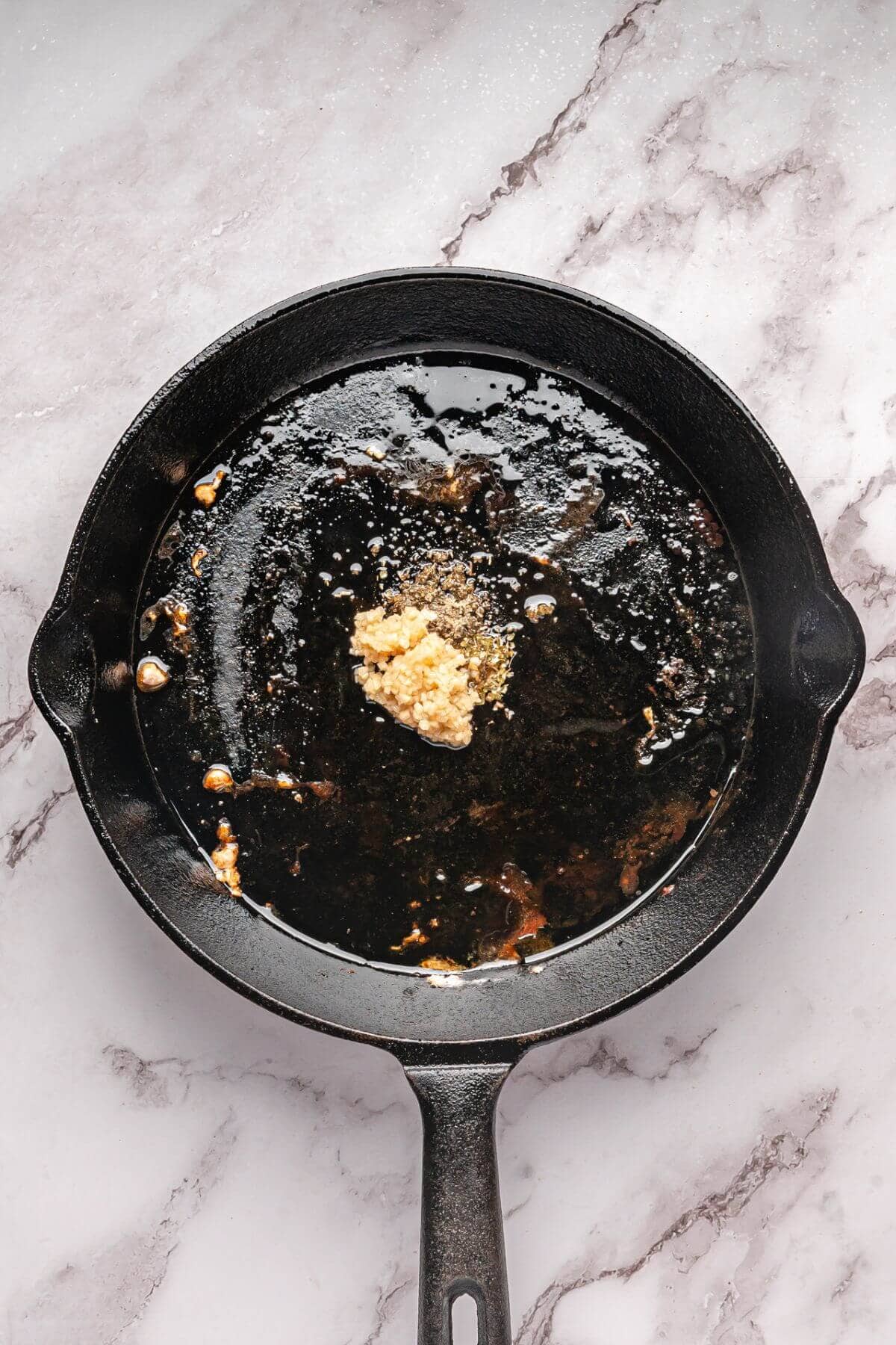 A cast iron skillet with oil residue and minced garlic inside, placed on a white marble surface.
