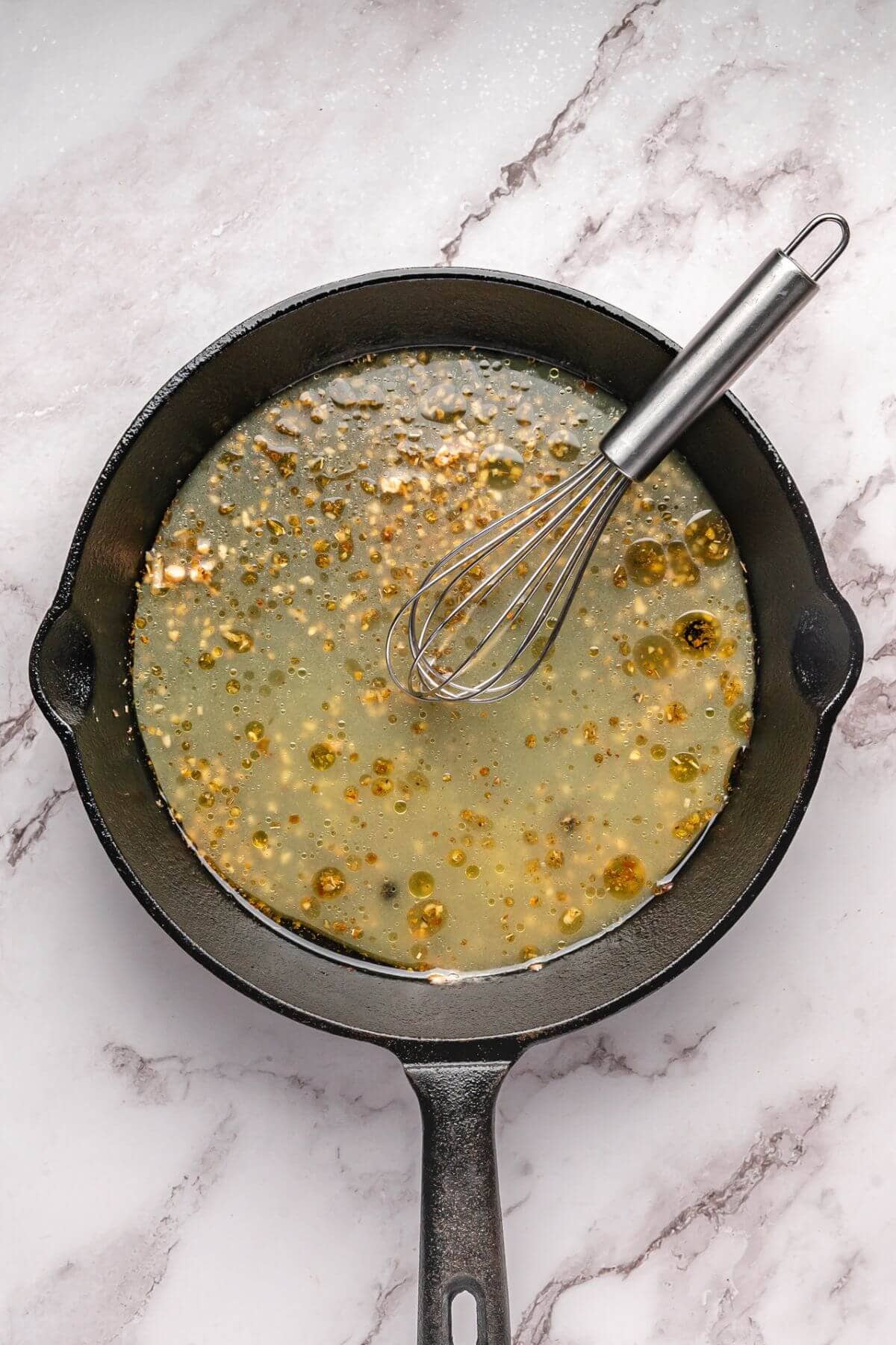 A black cast iron skillet with a metal whisk, containing a bubbling liquid sauce with visible bits, sits on a light marble surface.