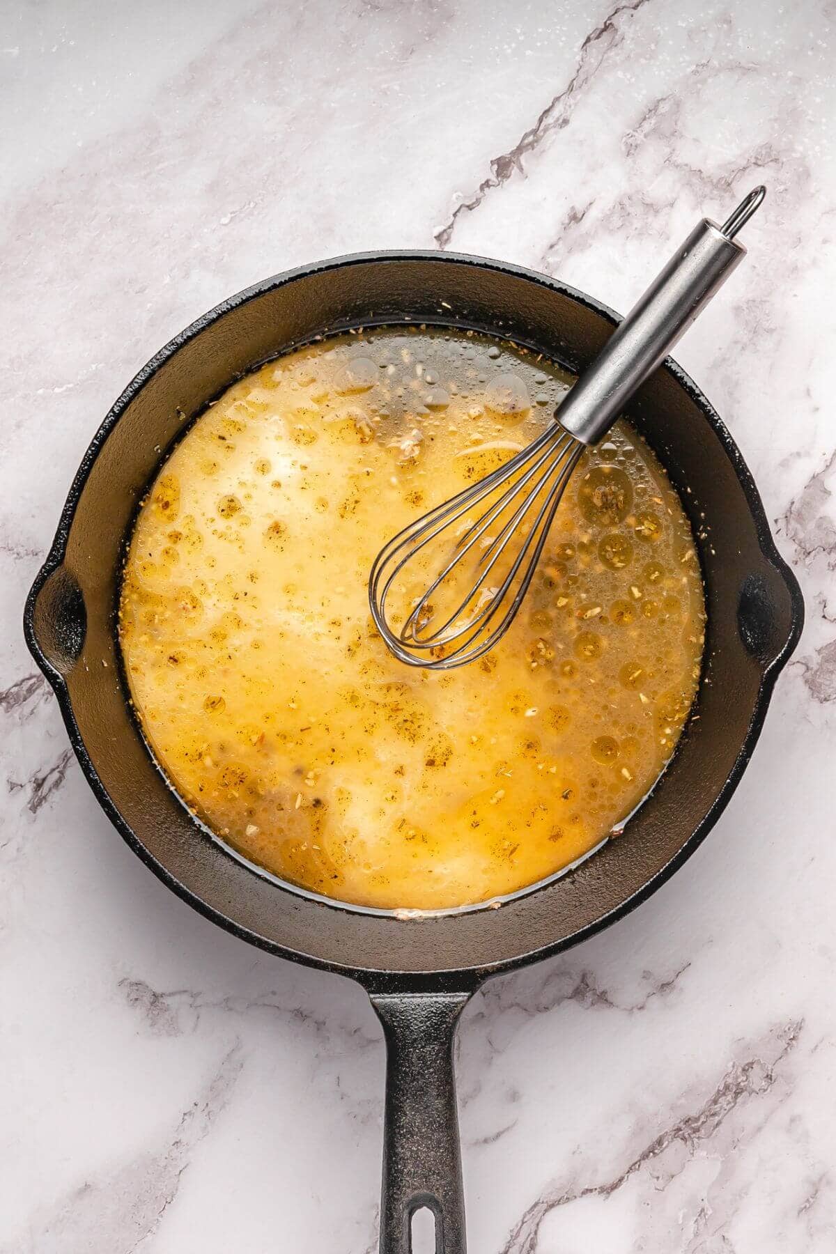 A cast iron skillet with a bubbling sauce being mixed by a metal whisk, set on a marble countertop.