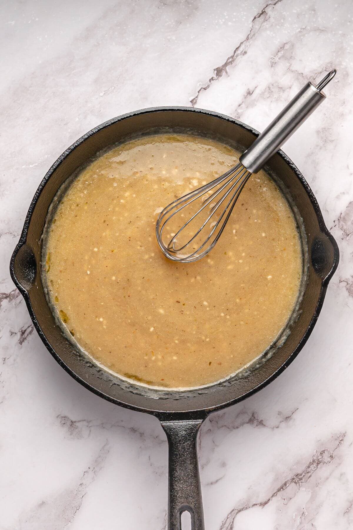 A cast iron skillet with light brown gravy and a metal whisk resting inside, placed on a marble surface.