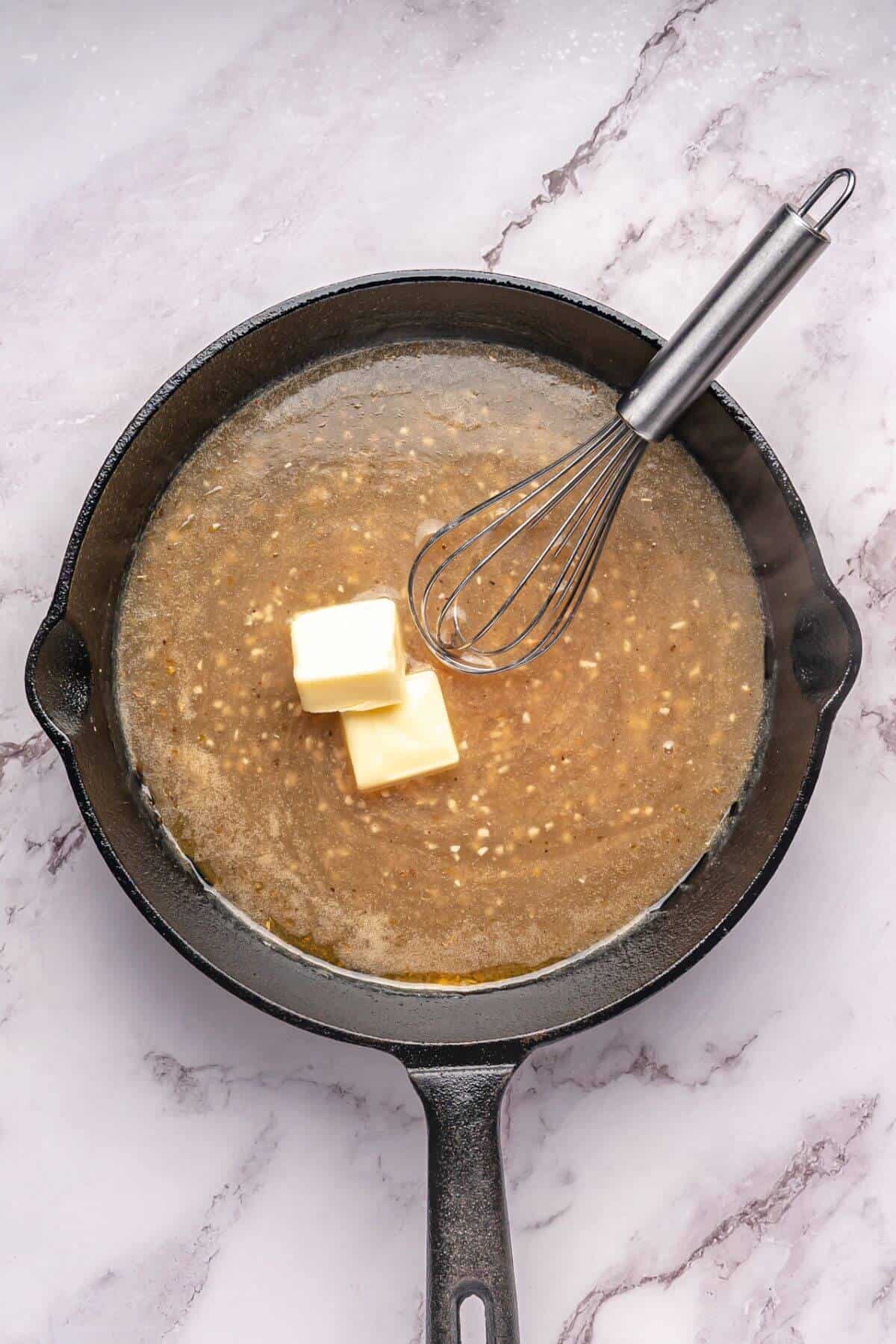 A cast iron skillet with melted mixture and two butter cubes, being whisked, is placed on a marble surface.