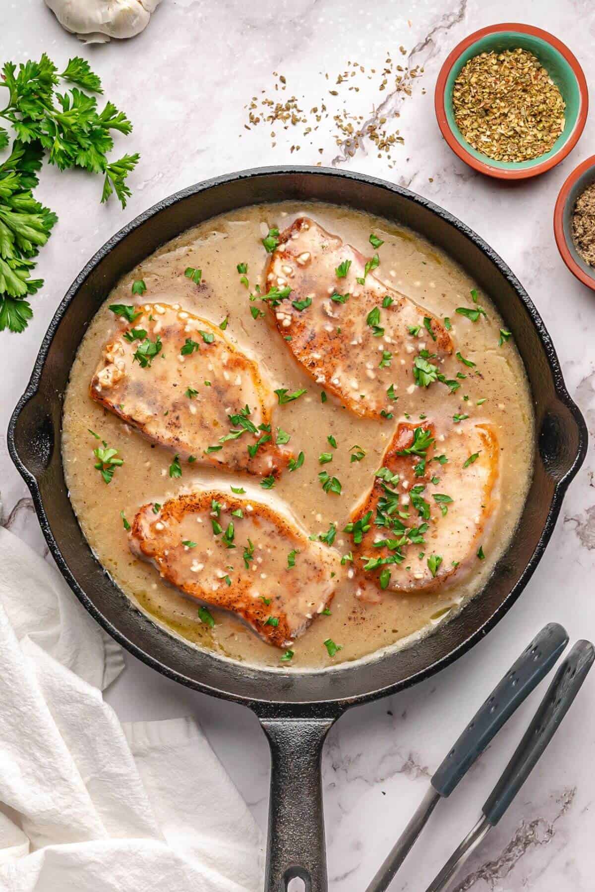 Four cooked pork chops in a creamy sauce, garnished with chopped herbs, in a cast iron skillet on a marble surface with seasonings and parsley nearby.