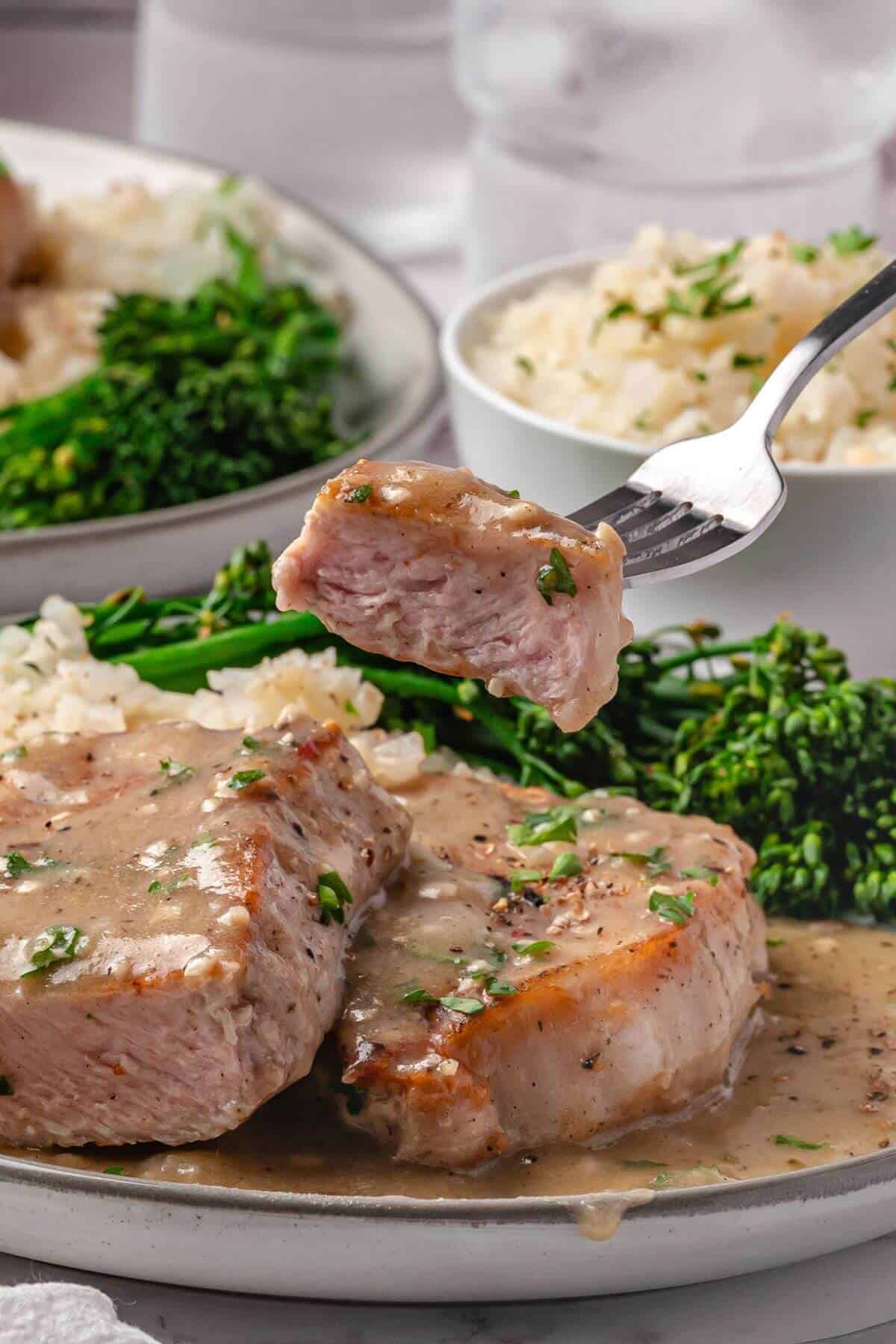 A fork holds a piece of pork chop with creamy sauce above a plate with more pork, broccolini, and rice in the background.