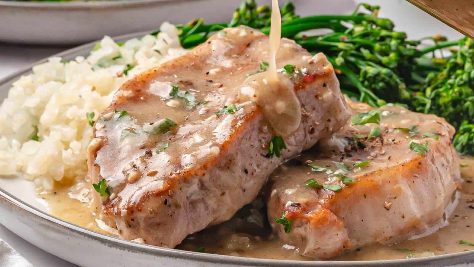 Two pork chops covered in creamy gravy on a plate, served with rice and steamed broccolini in the background.