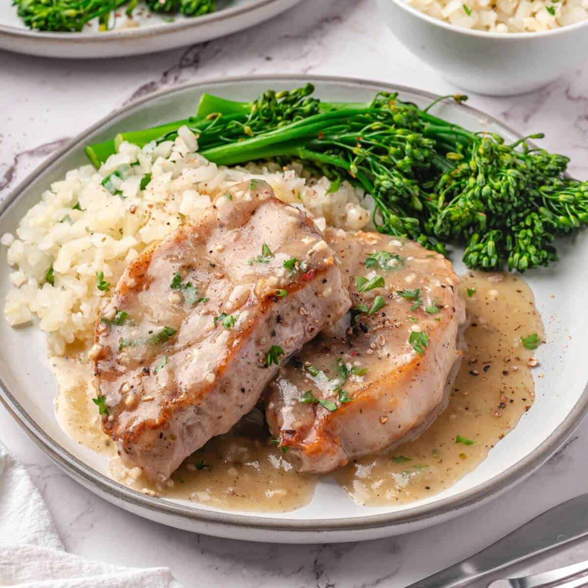 Two pork chops with gravy, served with white rice and steamed broccolini on a white plate.