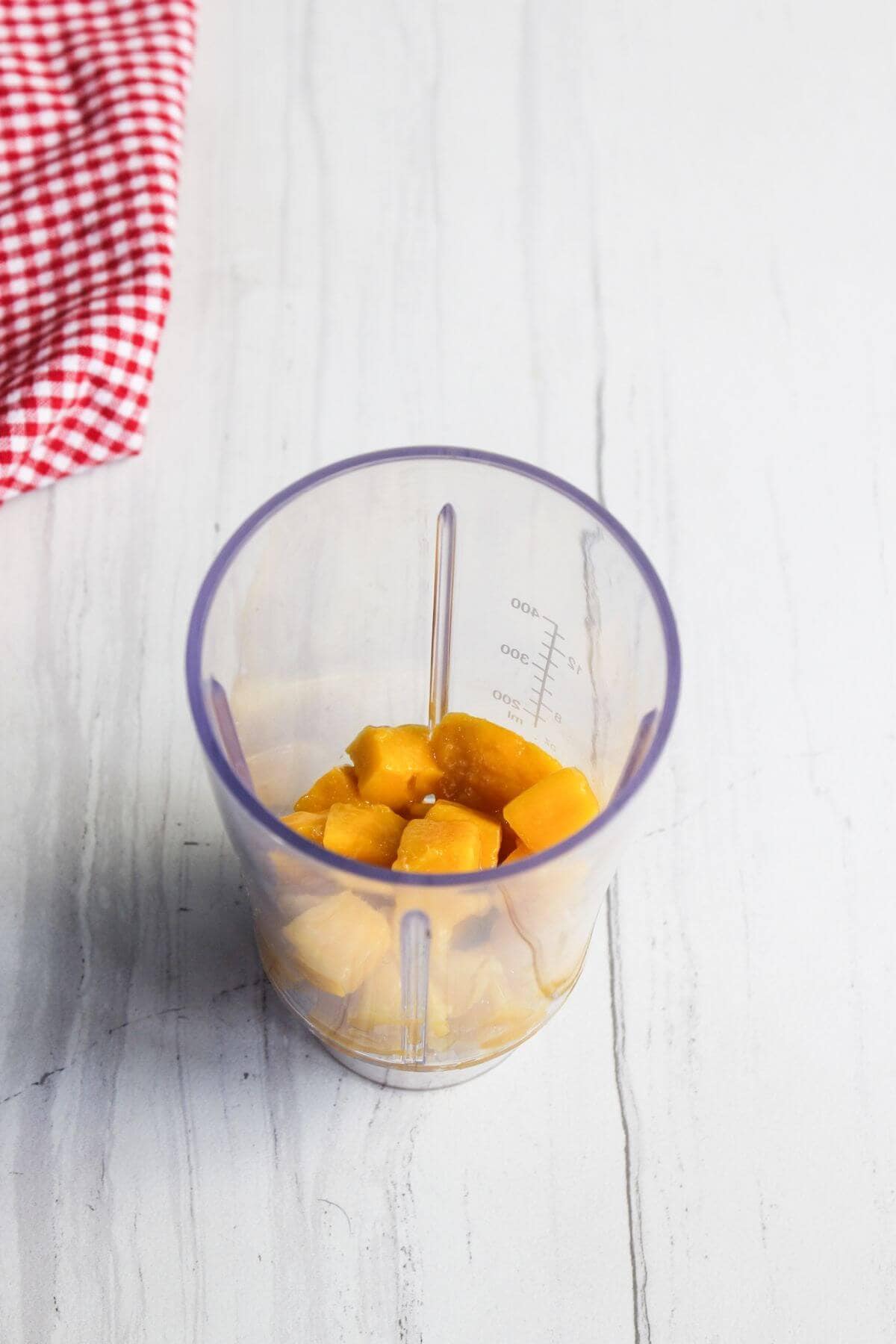Clear blender cup containing several cubes of mango on a light-colored surface, with a red and white checkered cloth partially visible in the upper left corner.