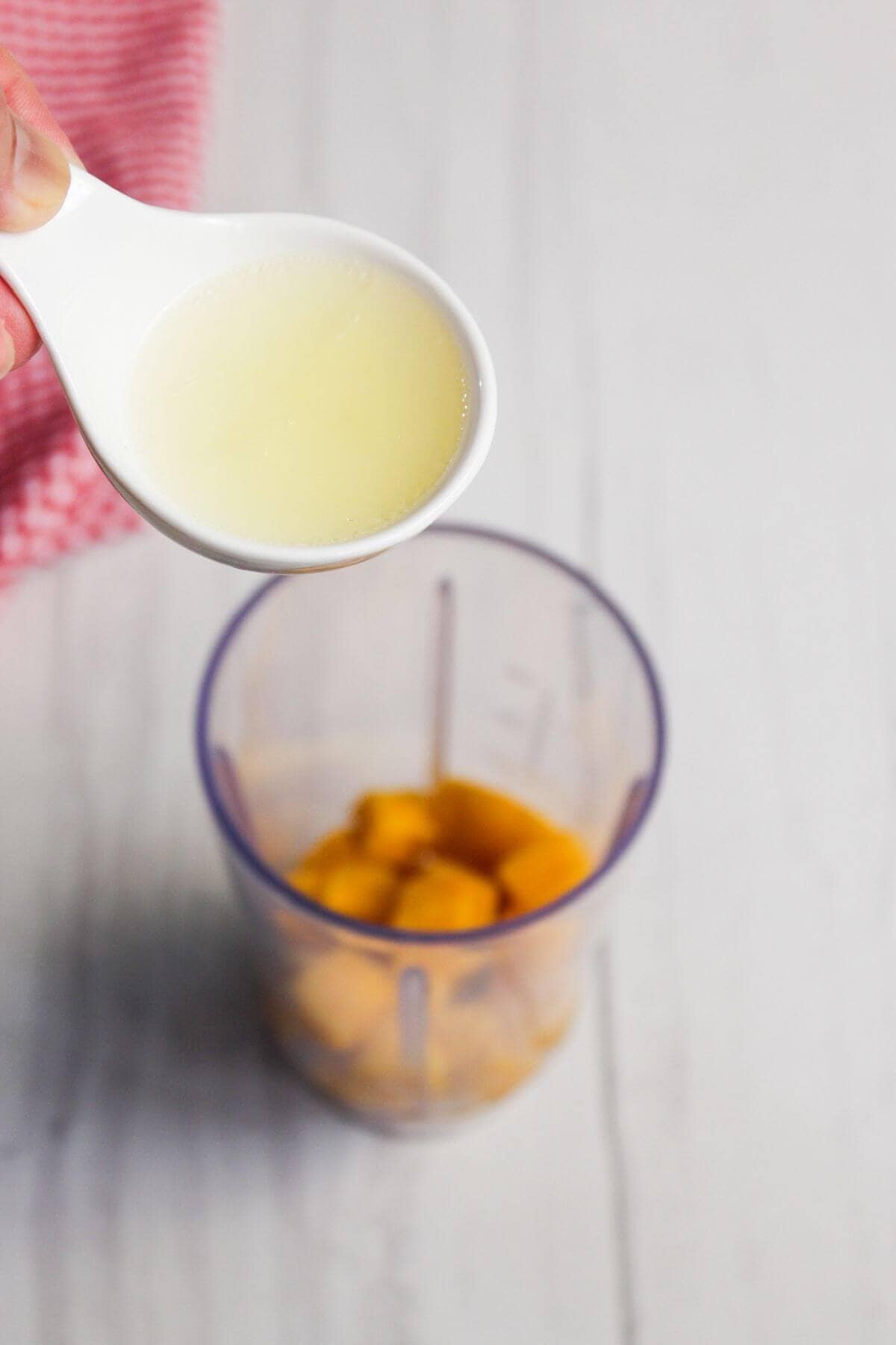 A hand holds a white spoon of lemon juice above a blender cup containing mango chunks on a light-colored surface.