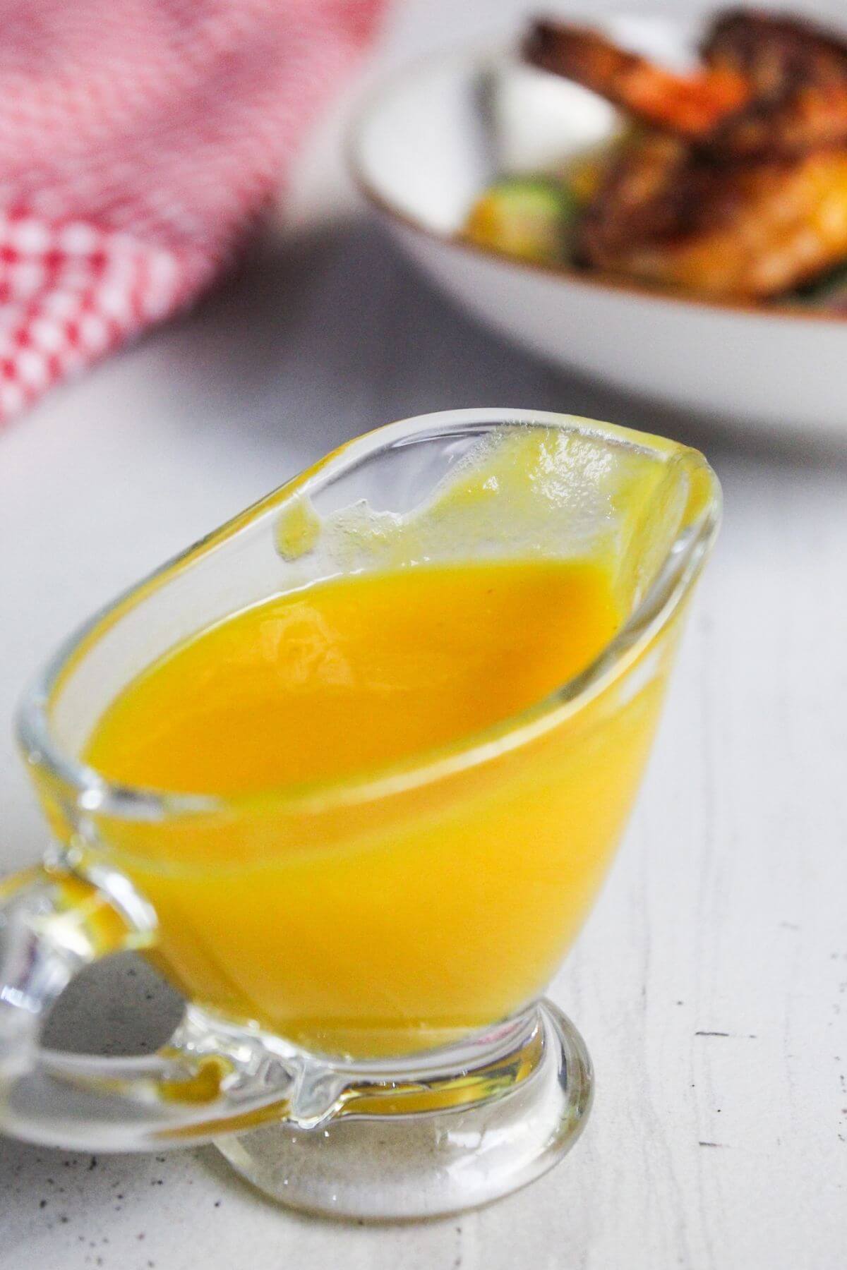 A glass gravy boat filled with bright yellow sauce sits on a white surface, with a plate of food blurred in the background.