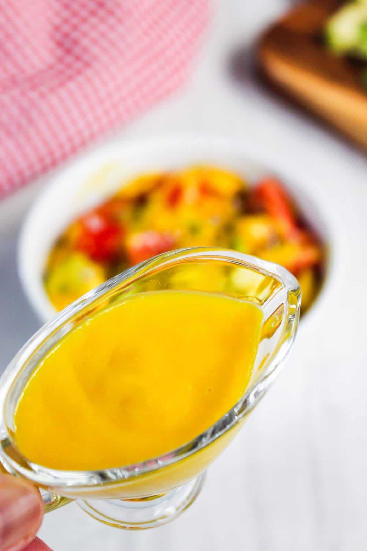 A close-up of a glass gravy boat filled with yellow sauce, held above a bowl of food containing vegetables.