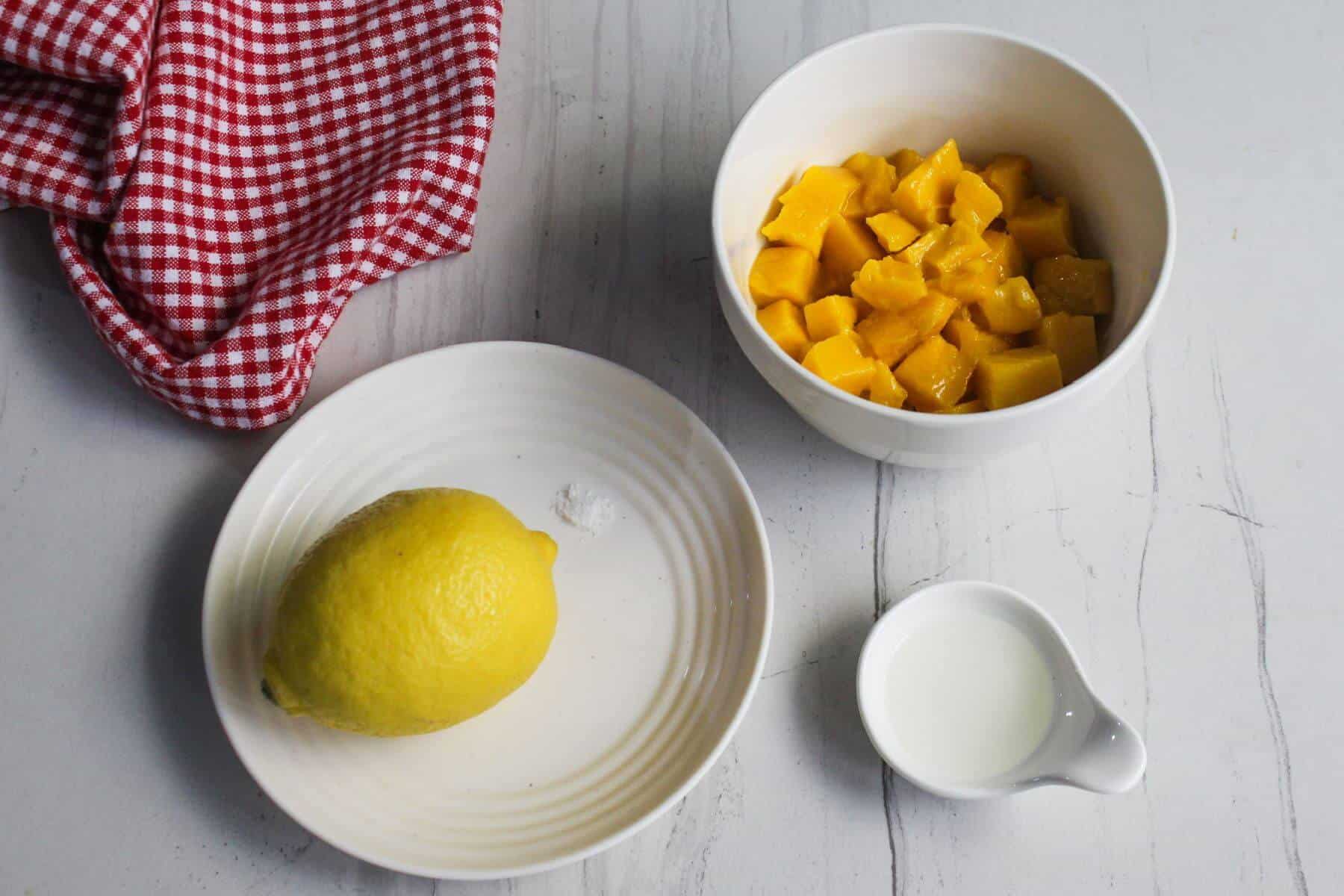 A whole lemon on a plate with a pinch of salt, a bowl of diced mangoes, a small jug of milk, and a red checkered cloth on a white surface.