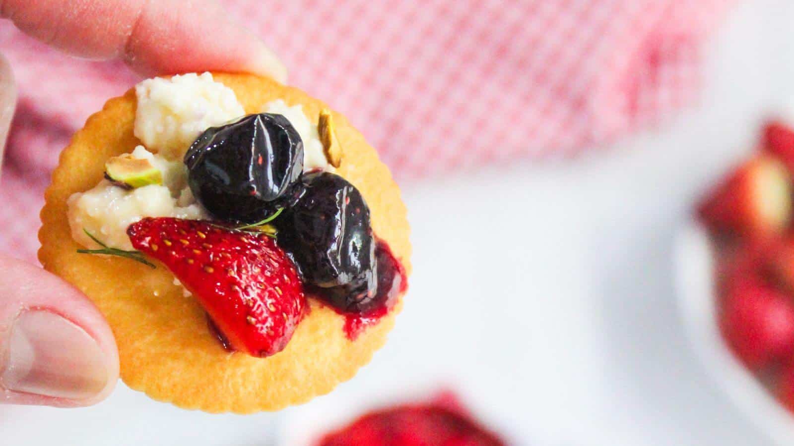 A hand holds a round cracker topped with white cheese, a strawberry slice, and two dark berries against a blurred background.