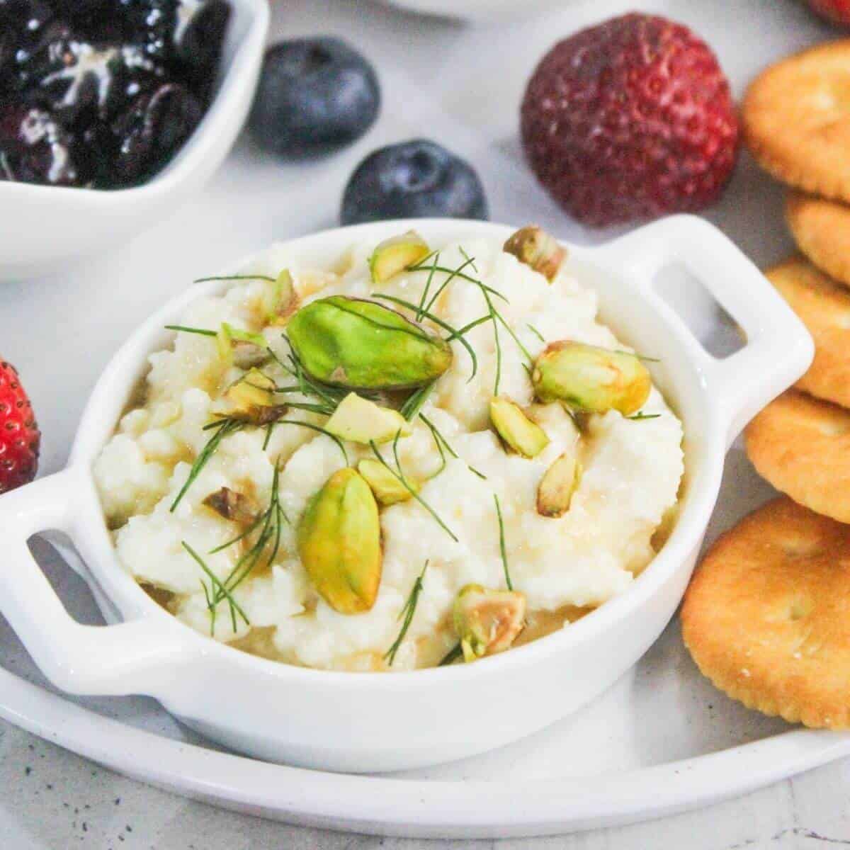 A small white dish of ricotta cheese topped with pistachios and herbs, surrounded by strawberries, blueberries, crackers, and a bowl of jam.