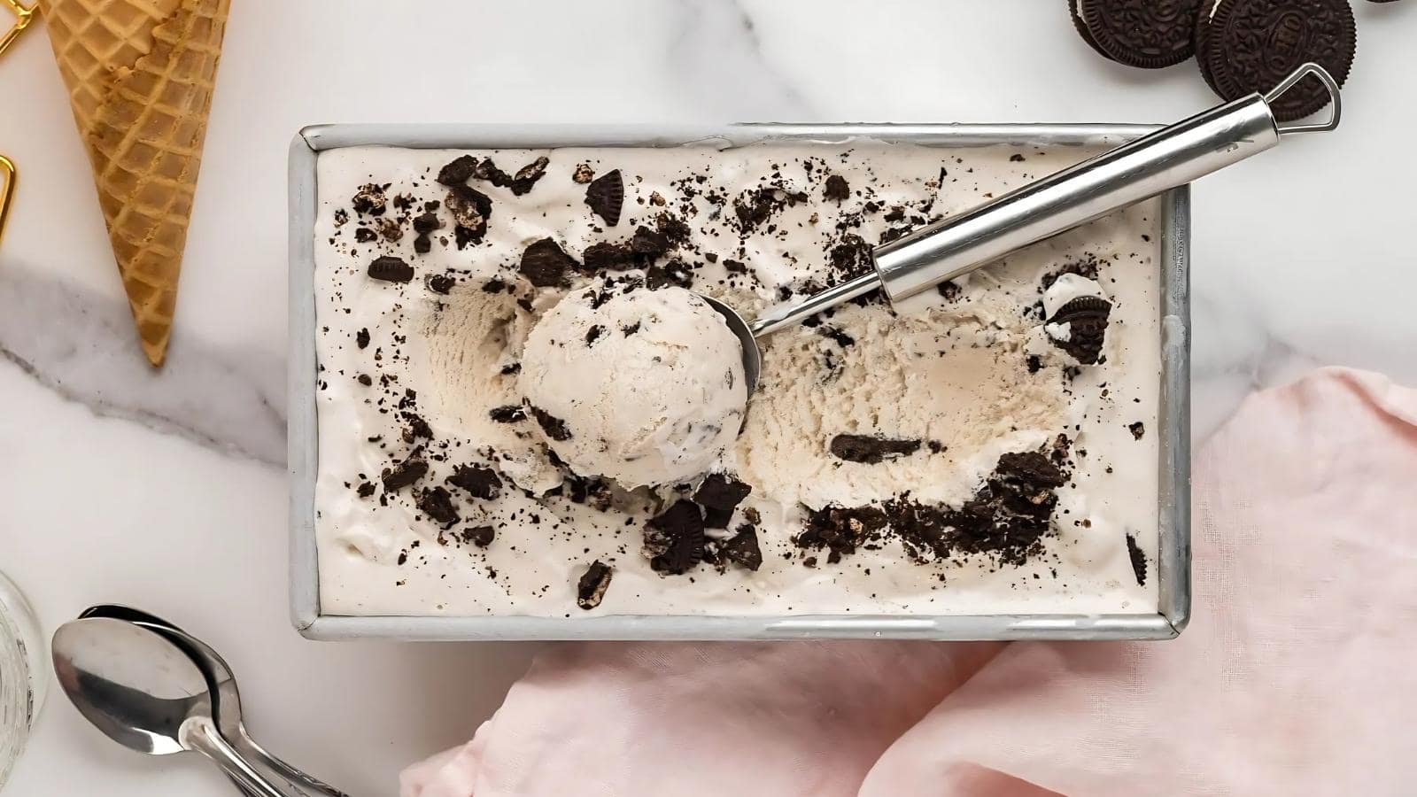 Rectangular metal container with cookies and cream ice cream, a scoop inside holding a ball of ice cream, crumbled cookies on top, a cone, and Oreo cookies nearby on a marble surface.