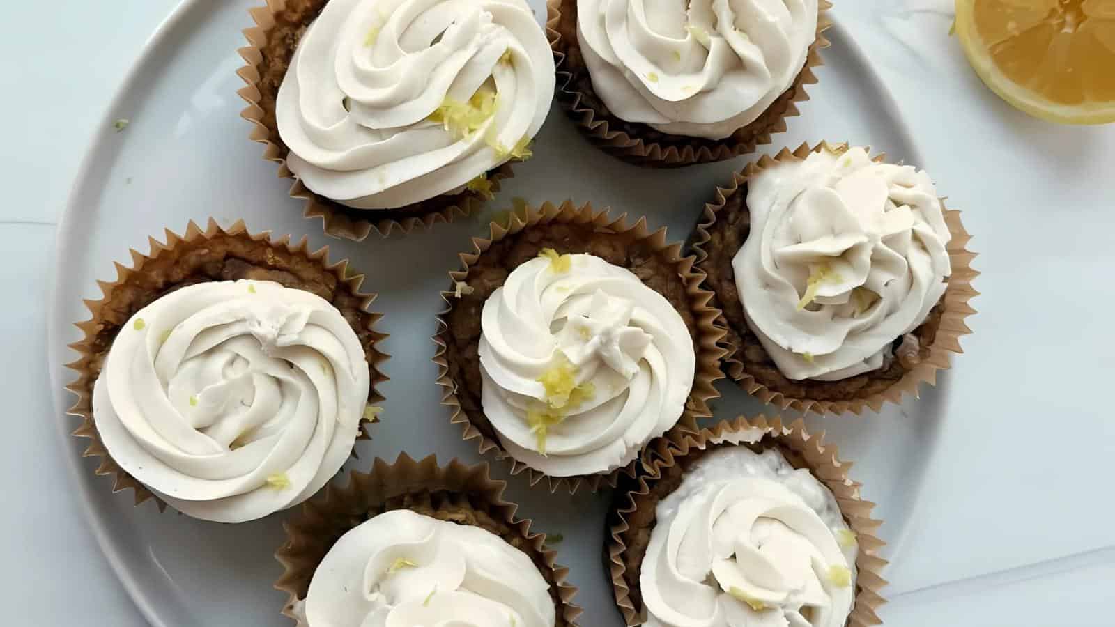 Seven cupcakes with white frosting and lemon zest are arranged on a white plate; a lemon slice is partially visible in the top right corner.