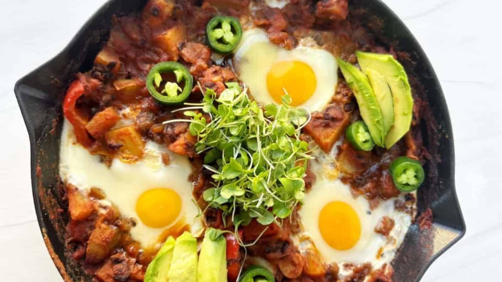 A skillet with three sunny-side-up eggs, tomato-based sauce, sliced avocado, jalapeño, and fresh greens on top.