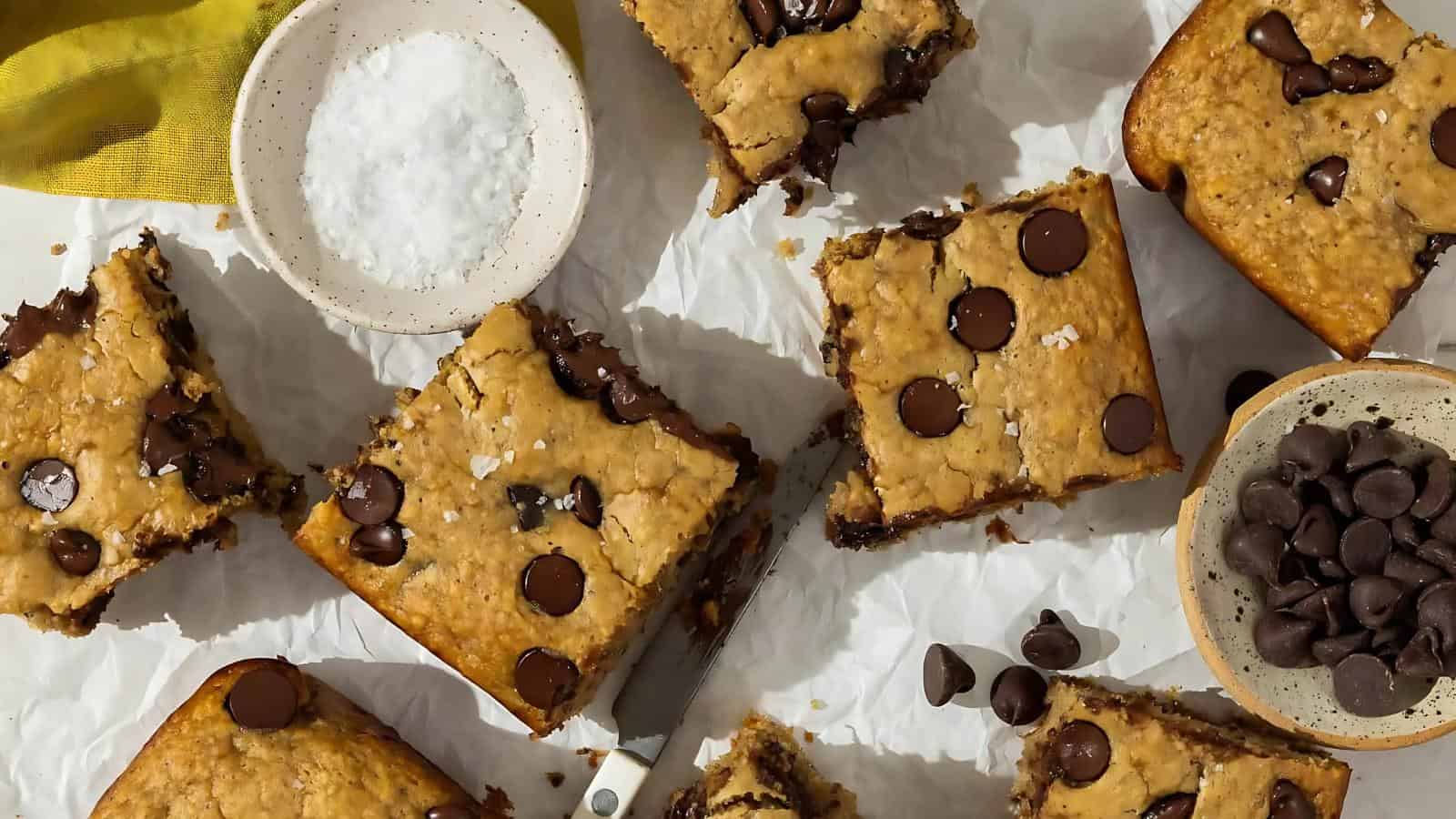 Several chocolate chip blondies are arranged on parchment paper, surrounded by a bowl of chocolate chips and a bowl of flaky sea salt. A knife rests among the cut blondies.