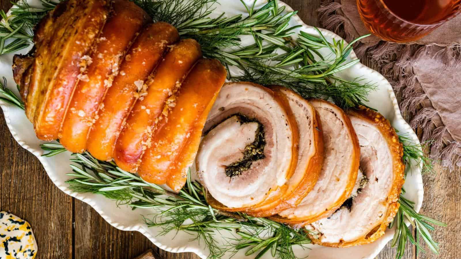 A rolled and roasted pork belly, partially sliced, is served on a white platter garnished with fresh rosemary and dill.