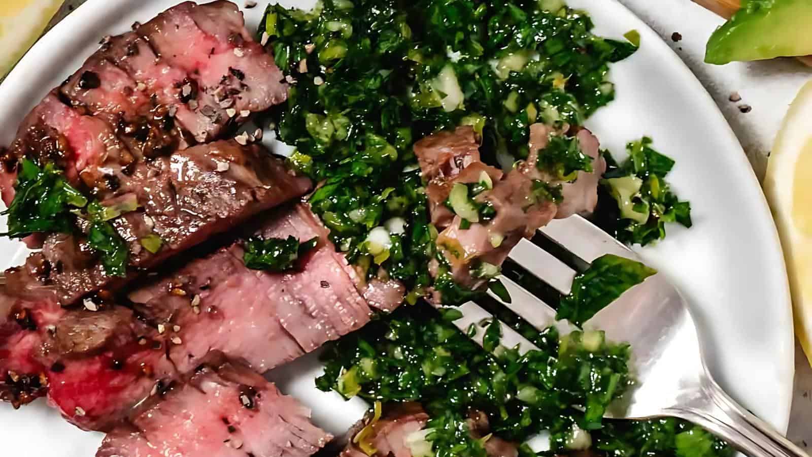 Slices of medium-rare steak with chimichurri sauce on a white plate, with a fork resting on the plate.