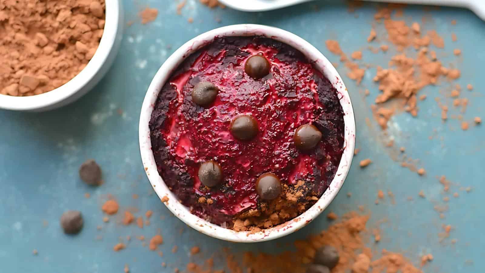 A small white ramekin filled with a baked dessert topped with berry sauce and chocolate pieces, surrounded by cocoa powder and chocolate chips on a blue surface.