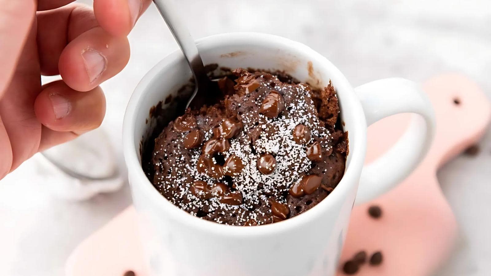 A hand holds a spoon about to scoop a chocolate chip mug cake topped with powdered sugar in a white mug.