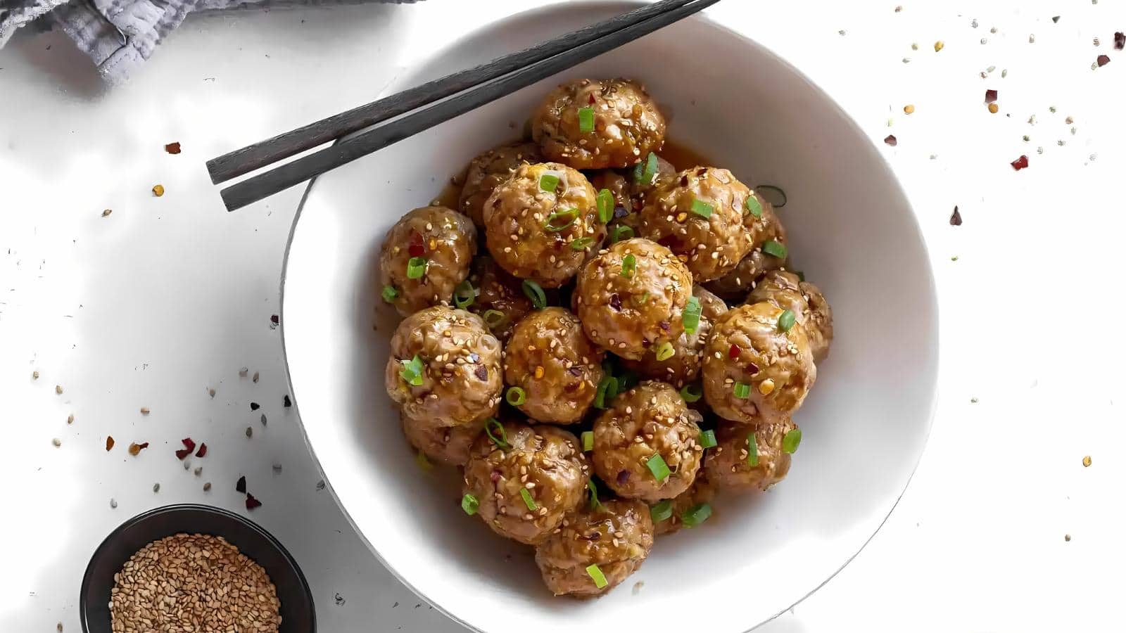 A white bowl filled with meatballs garnished with chopped green onions and sesame seeds, with a pair of chopsticks resting on top.