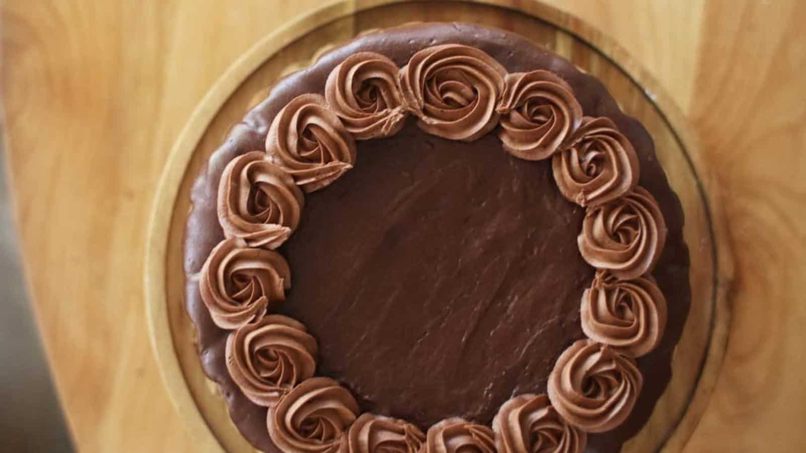 A chocolate cake with smooth frosting, decorated with a ring of chocolate rosettes around the edge, sits on a wooden surface.