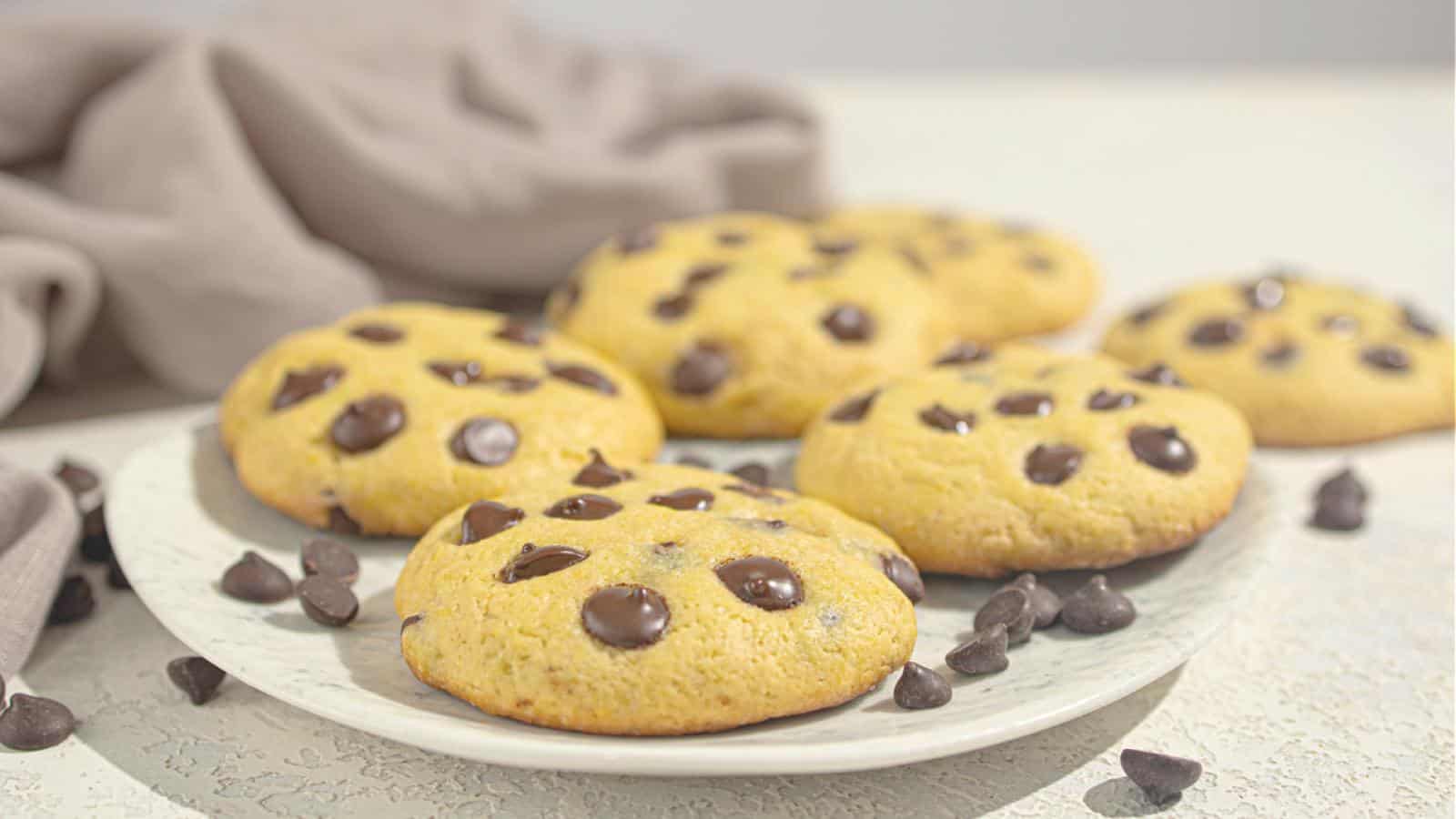 Six chocolate chip cookies are arranged on a white plate, with extra chocolate chips scattered on the table nearby. A beige cloth is in the background.