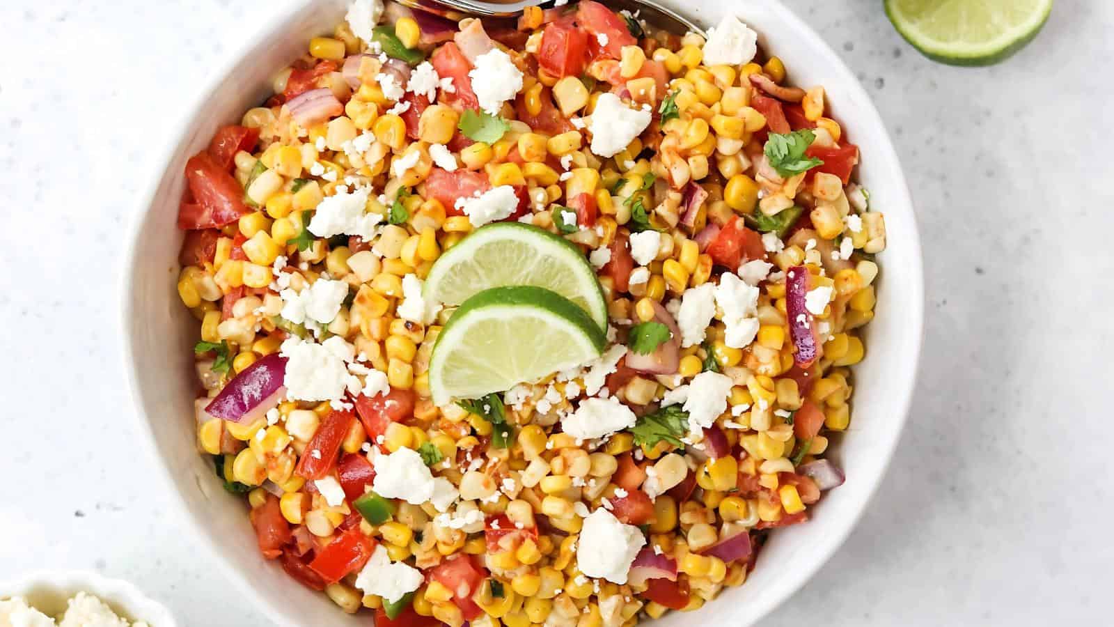A bowl of corn salad with diced tomatoes, red onion, cilantro, crumbled cheese, and lime slices on top.