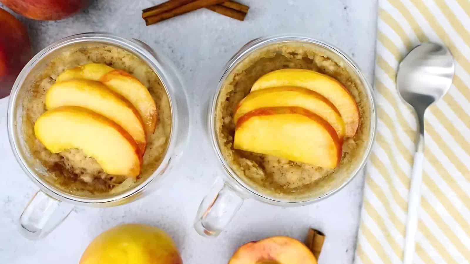 Two glass mugs filled with oatmeal, each topped with sliced peaches. A spoon, a striped napkin, cinnamon sticks, and whole peaches are arranged nearby on a light surface.