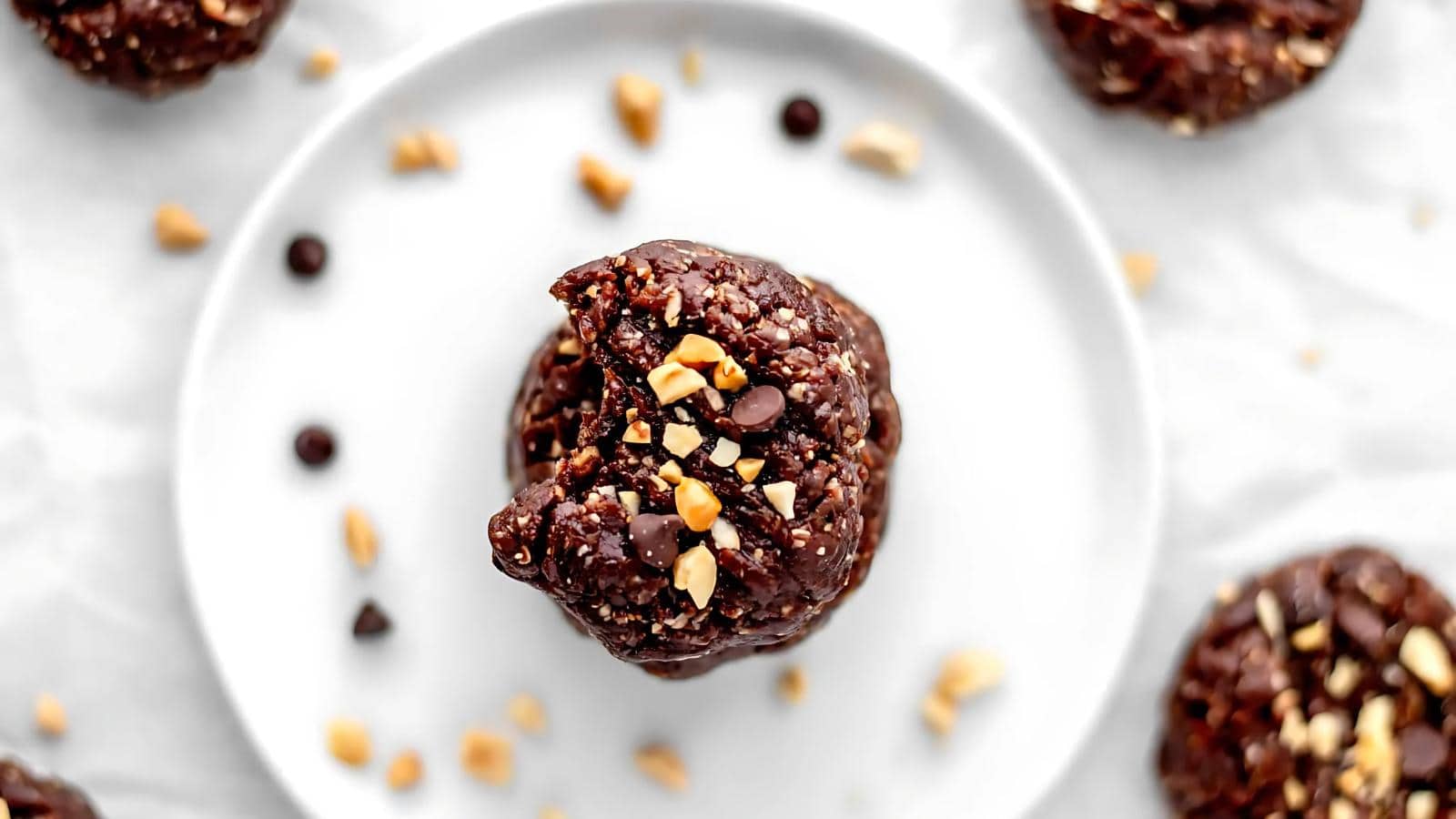 A close-up of a chocolate cookie topped with chopped peanuts and chocolate chips on a white plate, surrounded by more cookies and scattered toppings.