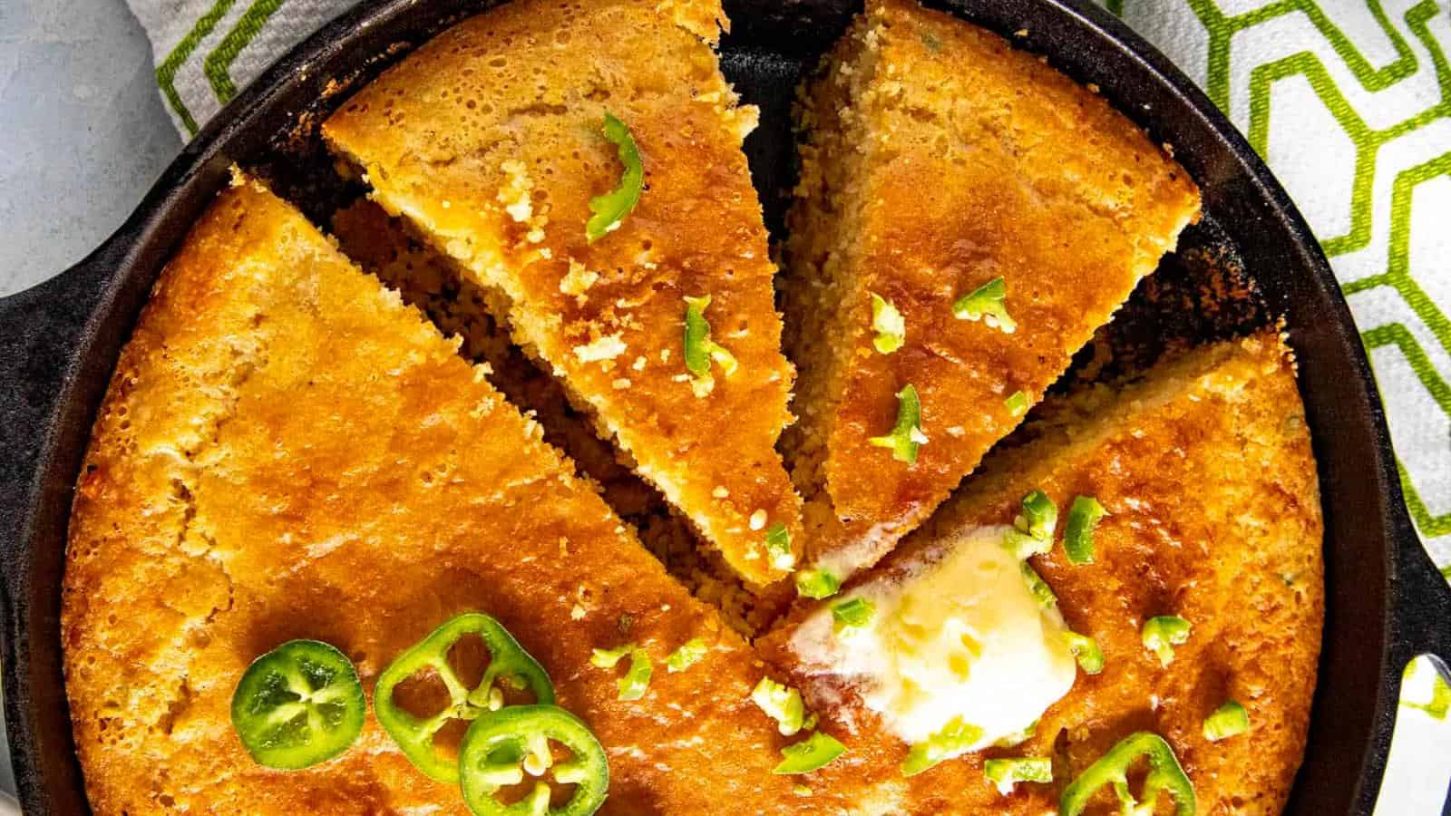 A skillet of cornbread cut into wedges, topped with sliced jalapeños and a dollop of melting butter.
