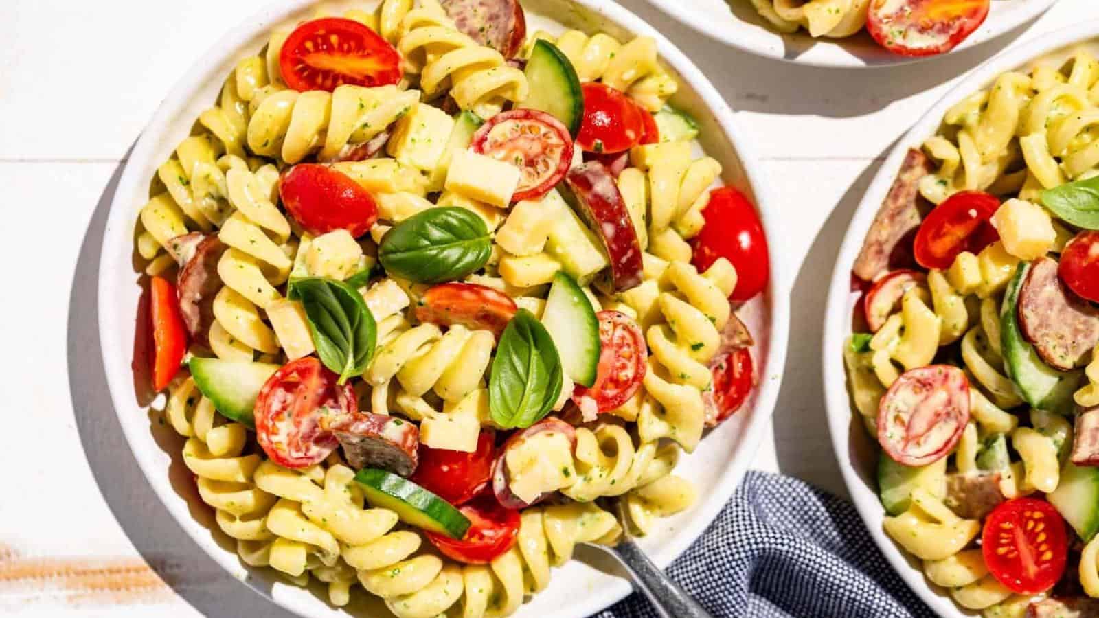 A bowl of rotini pasta salad with cherry tomatoes, cucumber slices, cheese cubes, sausage pieces, and fresh basil leaves, with a fork and a napkin beside the bowl.
