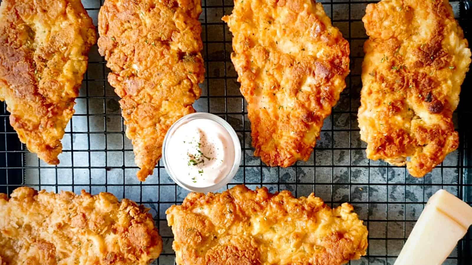 Six pieces of fried chicken on a cooling rack with a small bowl of dipping sauce and a wedge of cheese beside them.