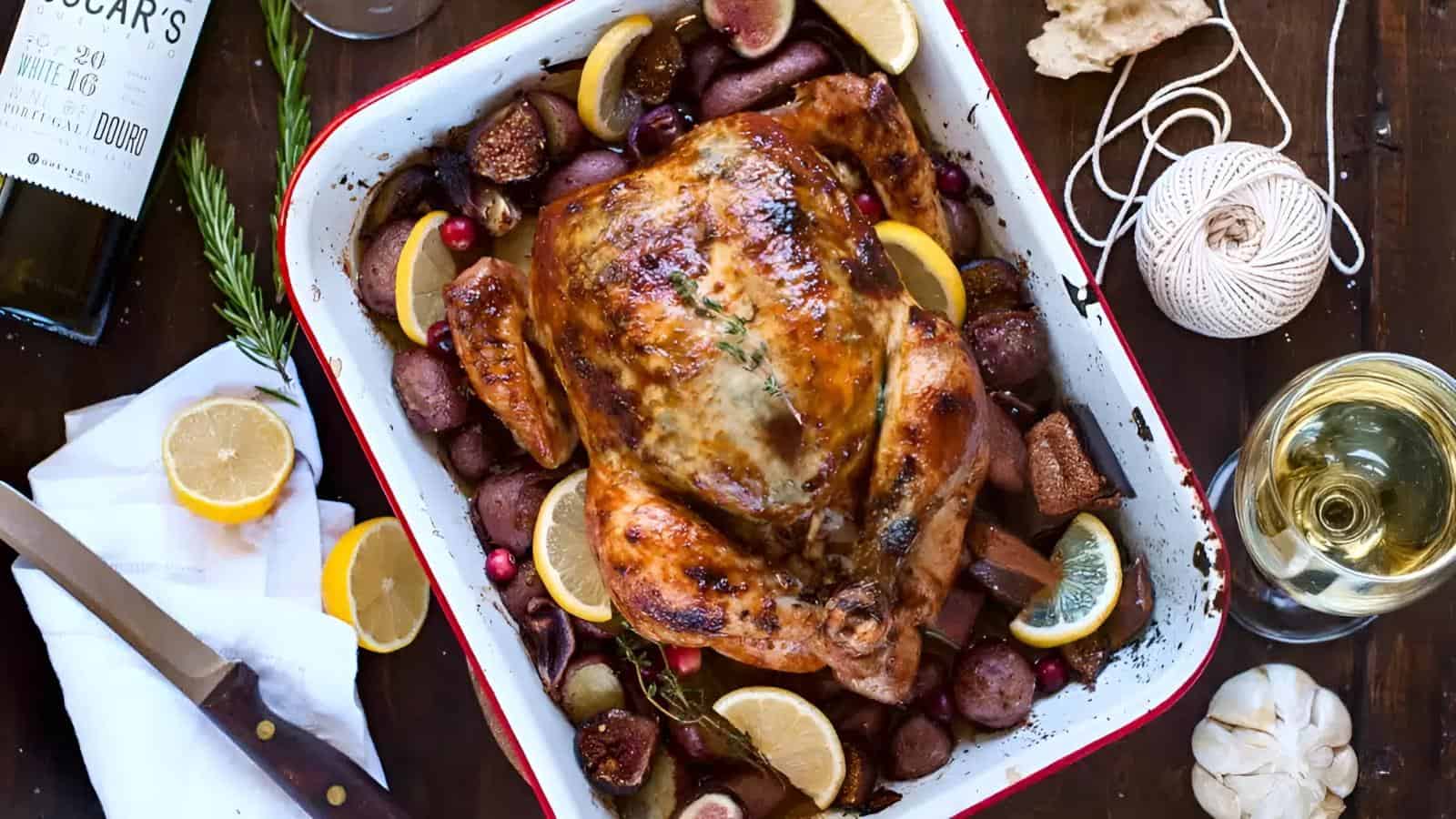 Roast chicken in a baking dish surrounded by lemon slices, figs, red potatoes, and herbs on a wooden table with wine and kitchen utensils nearby.