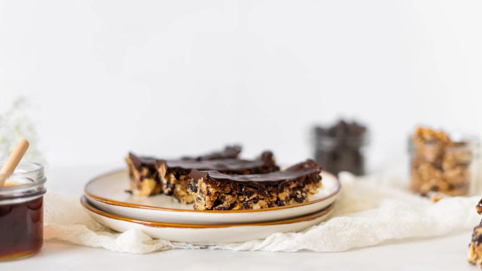 Three chocolate-topped dessert bars are stacked on a plate, with jars of honey and nuts visible in the background on a white surface.