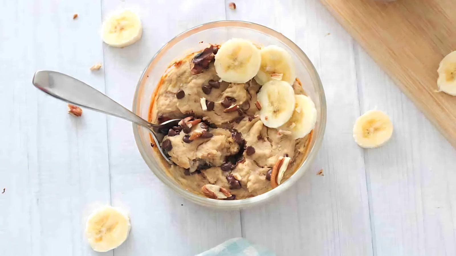 A glass bowl of oatmeal topped with banana slices, nuts, and chocolate chips, with a spoon inside, on a light wooden surface.