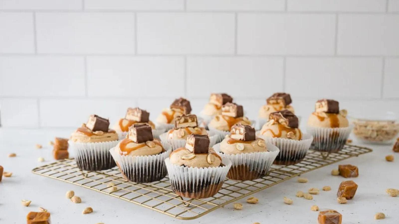 A cooling rack with cupcakes topped with caramel frosting, chopped nuts, and pieces of chocolate bars, with scattered peanuts and caramel pieces on a white surface.