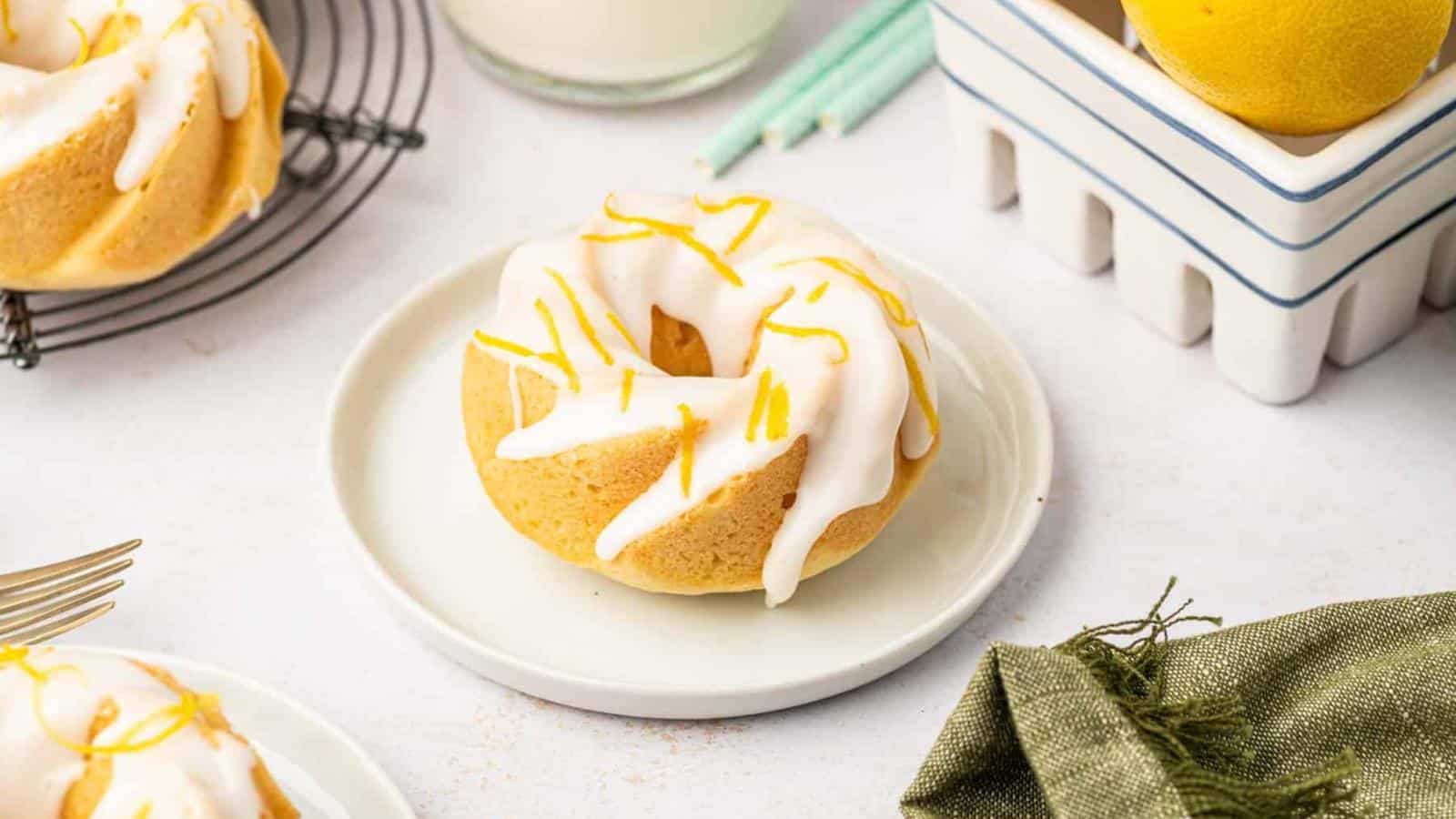 A small Bundt cake with white icing and lemon zest sits on a white plate, with lemons, a glass of milk, and candles in the background.