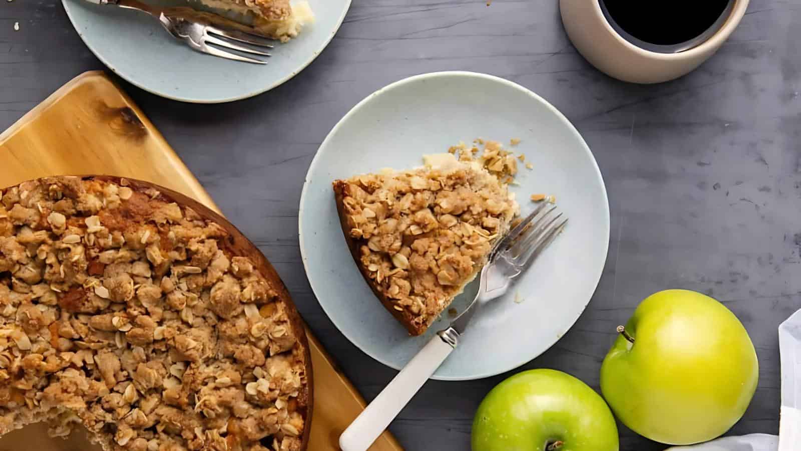 A slice of crumb-topped apple cake on a plate with a fork and knife, next to the whole cake, two green apples, and a cup of coffee on a gray surface.