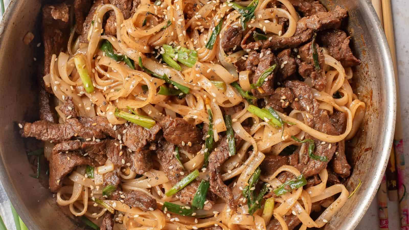 A skillet filled with stir-fried beef, flat rice noodles, green onions, and sesame seeds.