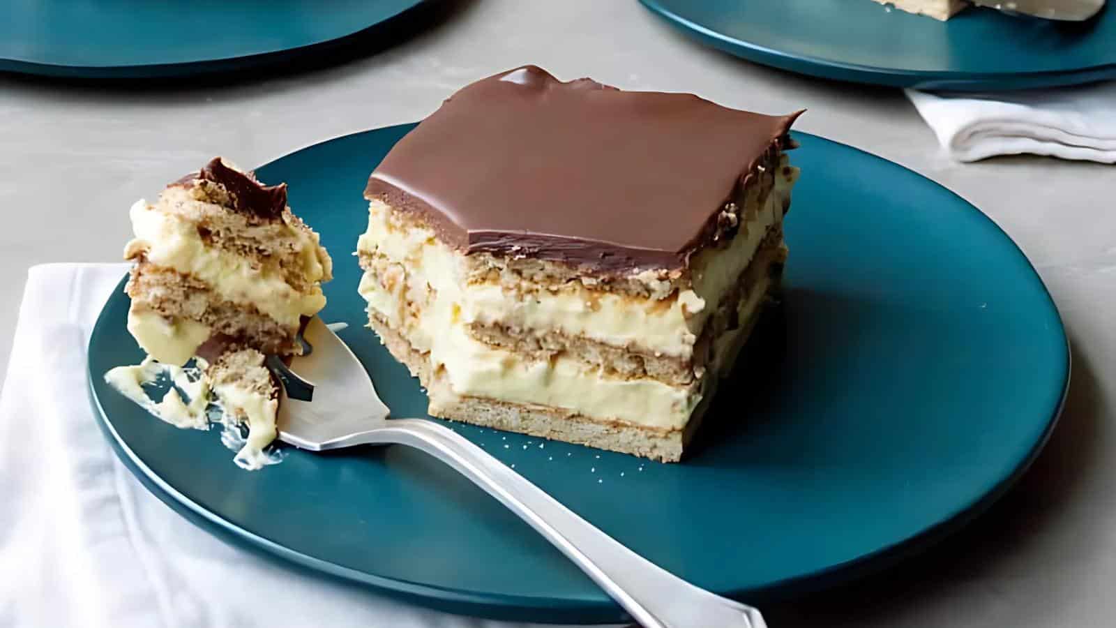 A square slice of layered dessert with chocolate topping, creamy filling, and cookie layers sits on a teal plate next to a fork with a bite taken.