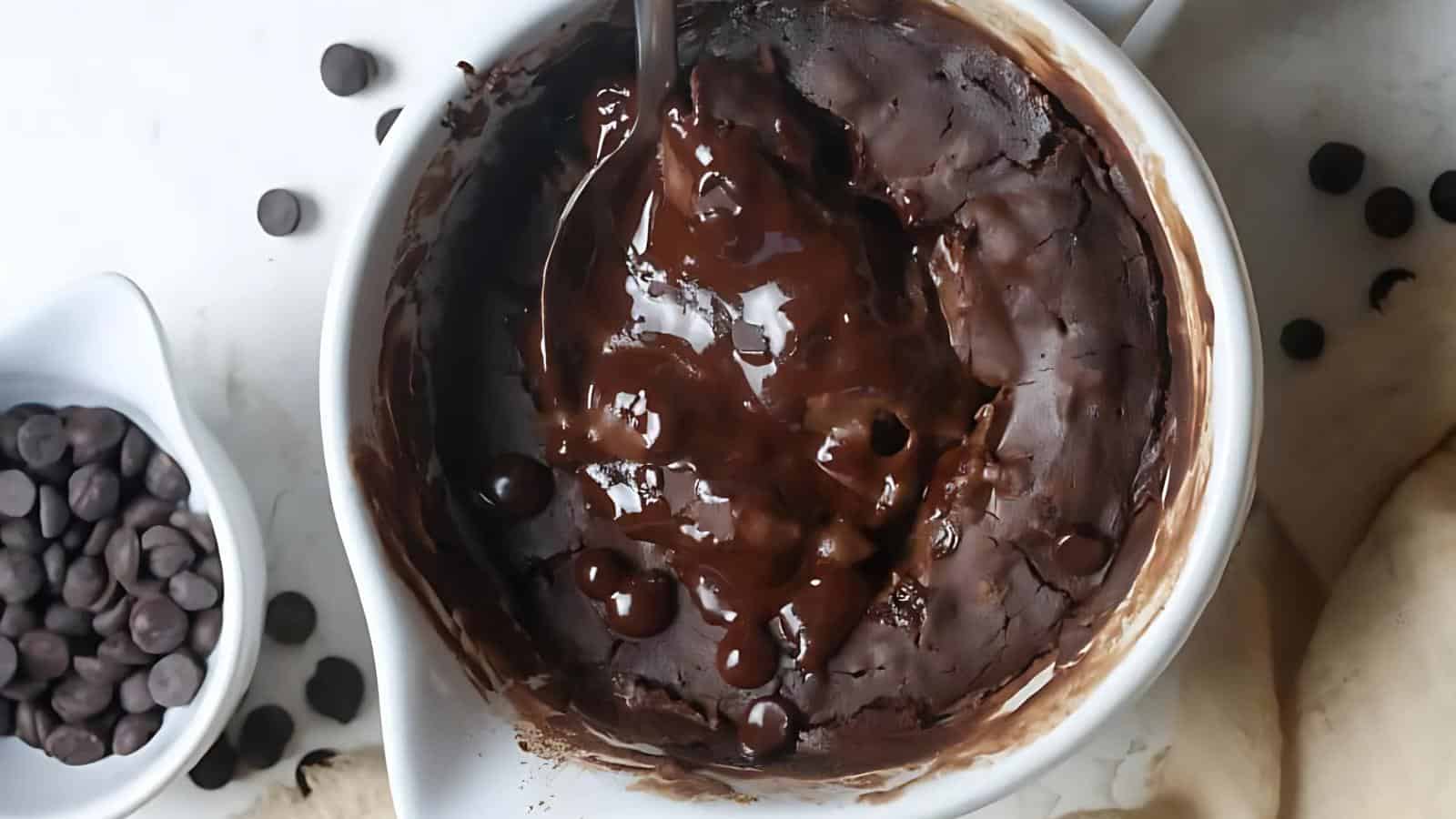 A chocolate mug cake with melted chocolate chips sits in a white mug, with a spoon inside. A small bowl of chocolate chips is next to the mug on a white surface.