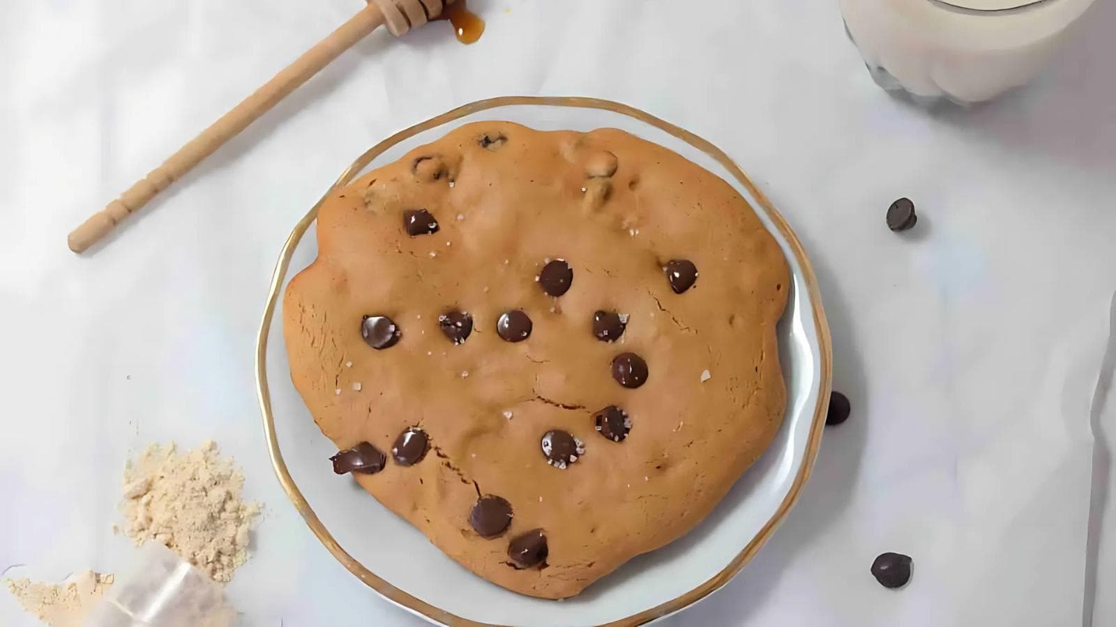 A large chocolate chip cookie on a white plate, surrounded by chocolate chips, flour, a honey dipper with honey, and a glass of milk on a white surface.