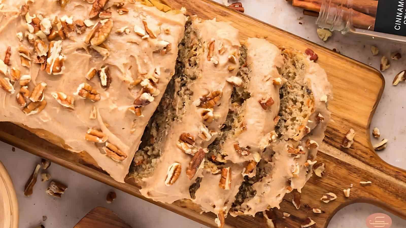 A loaf of frosted bread topped with chopped pecans is partially sliced on a wooden cutting board, with a jar of cinnamon sticks nearby.