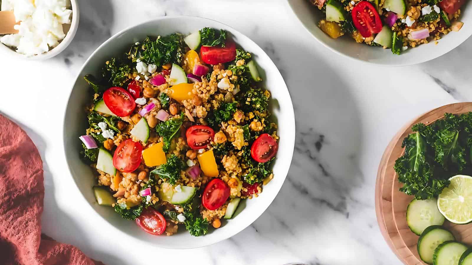 A bowl of salad with cherry tomatoes, kale, chopped cucumbers, yellow bell peppers, red onions, and grains on a marble surface, surrounded by salad ingredients.