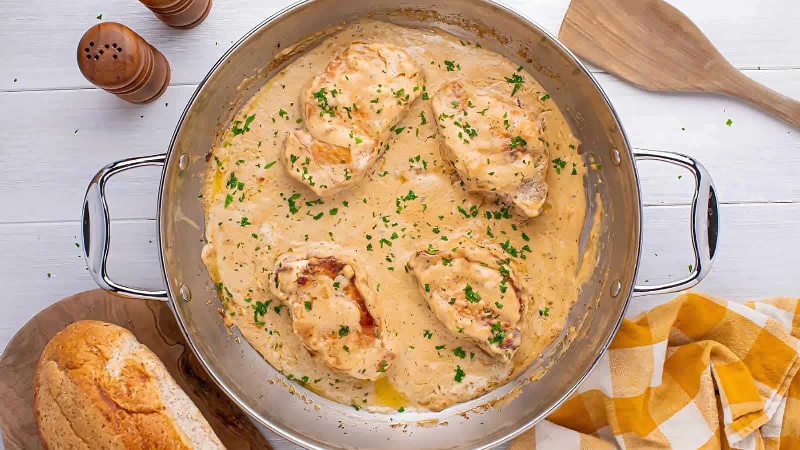 Four cooked chicken breasts in creamy sauce, garnished with chopped parsley, in a stainless steel skillet on a white table with bread, salt and pepper shakers, and a yellow checked napkin nearby.