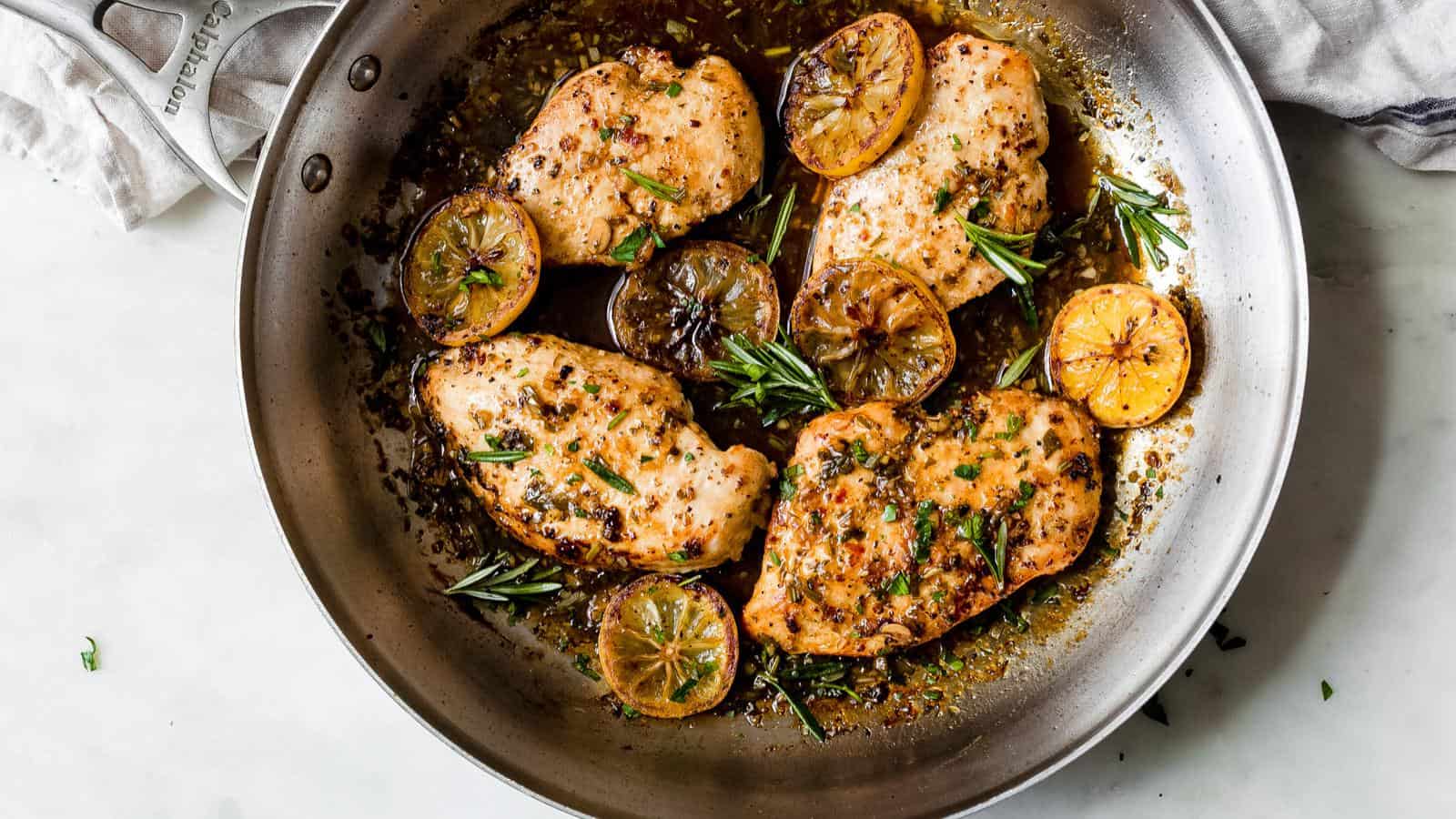 A stainless steel pan with cooked chicken breasts, sliced browned lemons, fresh rosemary, and herbs in a savory sauce.