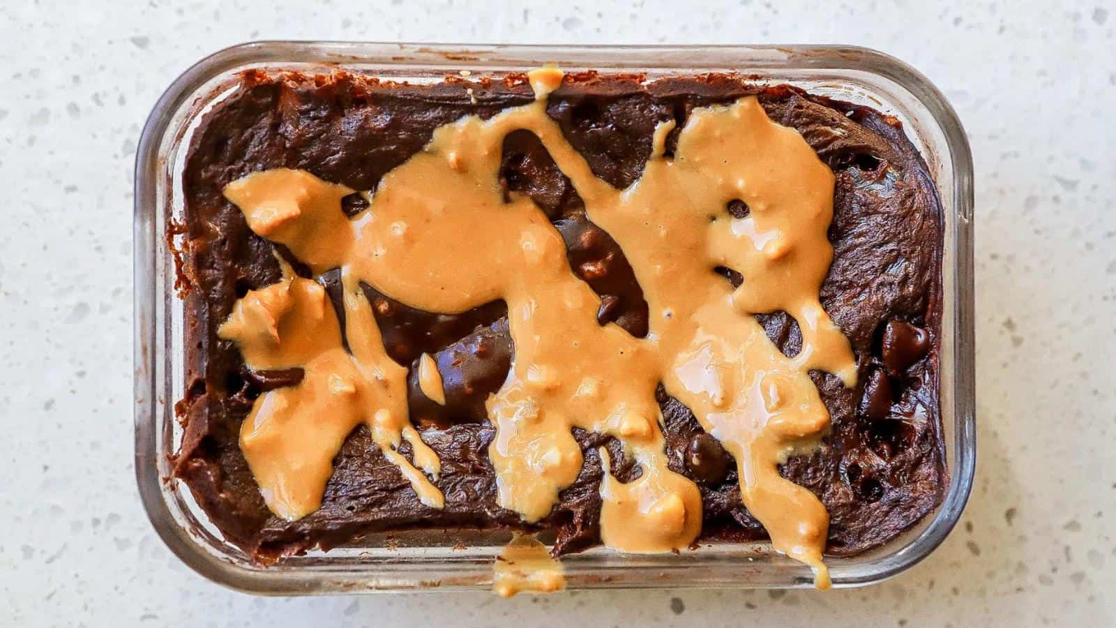 A glass dish filled with chocolate brownie topped with swirls of melted peanut butter, set on a light-colored countertop.